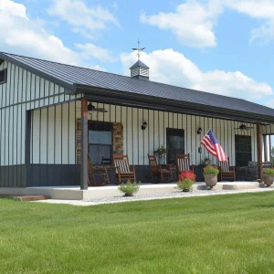 Beautiful 30x40 Barndominium on a grassy lawn. With flowers, rocking chairs and a flag at the front overhang before the home entrance.